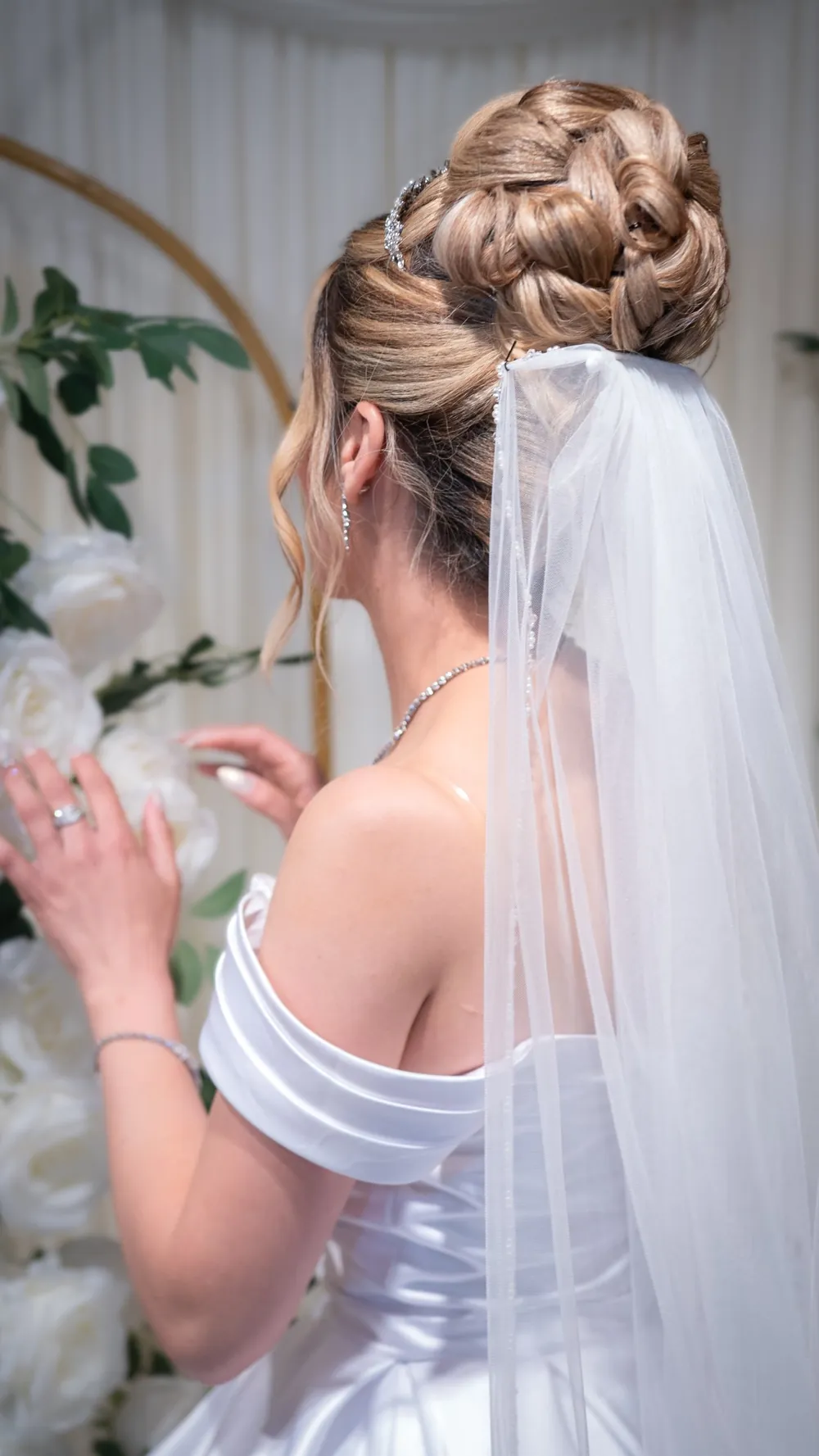 Bride in an off-shoulder satin gown and beaded veil, seen from behind beside white roses