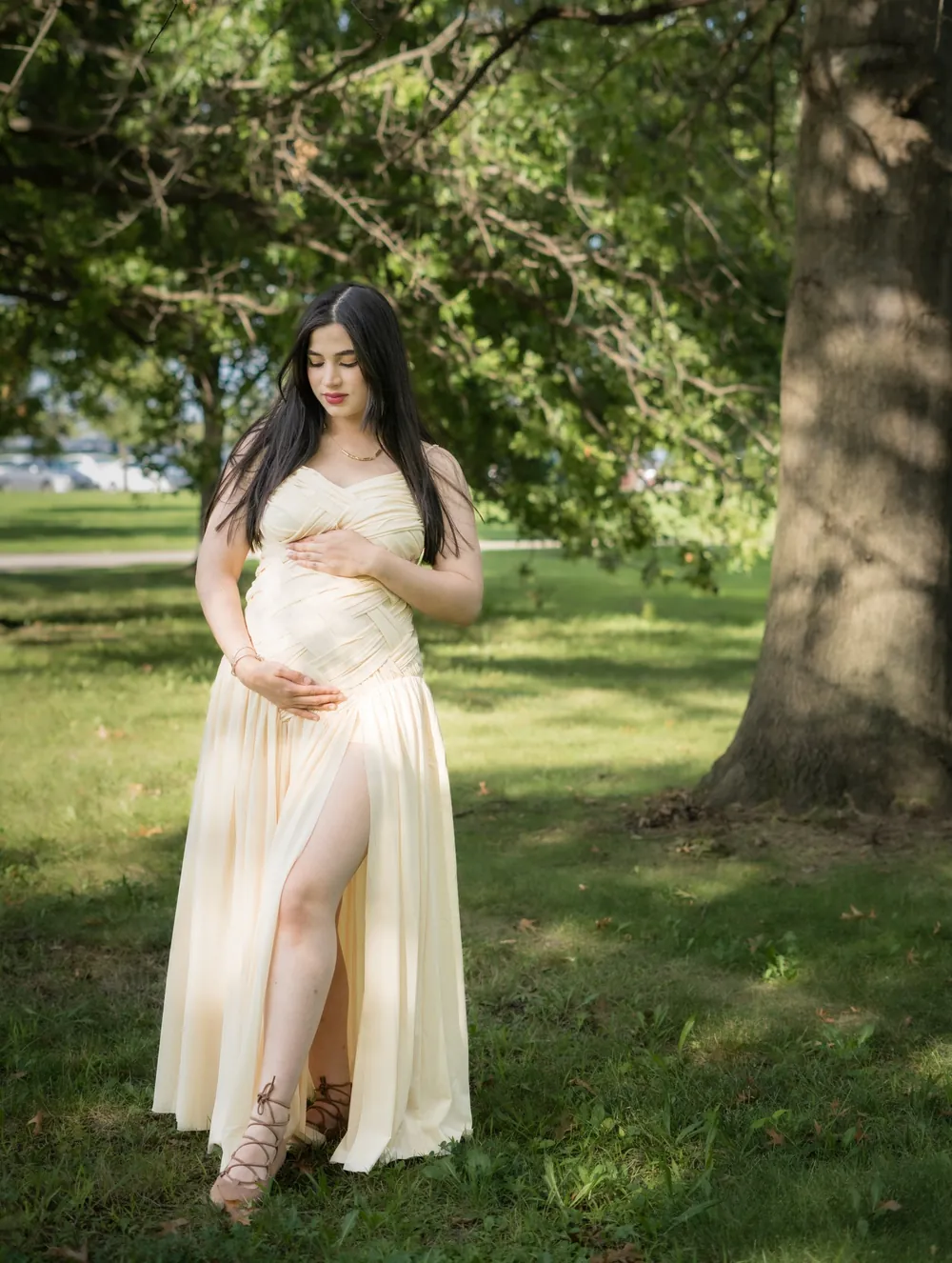 Expecting mother in a cream gown holding her belly beneath a tree in a Philadelphia park